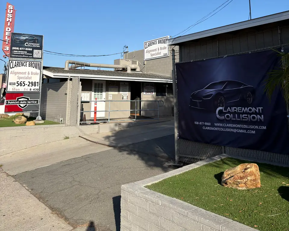 Street view of the Clairemont Collision driveway entrance on Armour St in San Diego, with the navy Clairemont Collision banner on the fence and neighboring business signs along the street